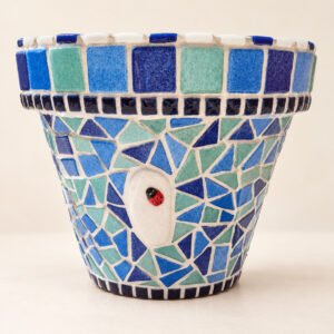 Pot de fleurs en mosaïque Azur vu de face, dans des tons bleus et vert d’eau, avec une coccinelle sur un galet blanc.